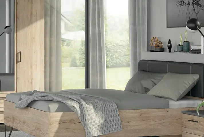 Doppelbett aus Holz mit grauer Bettwäsche und Decke, dahinter bodentiefe Fenster mit Vorhängen und Blick in den Garten, rechts Ablage mit Deko.